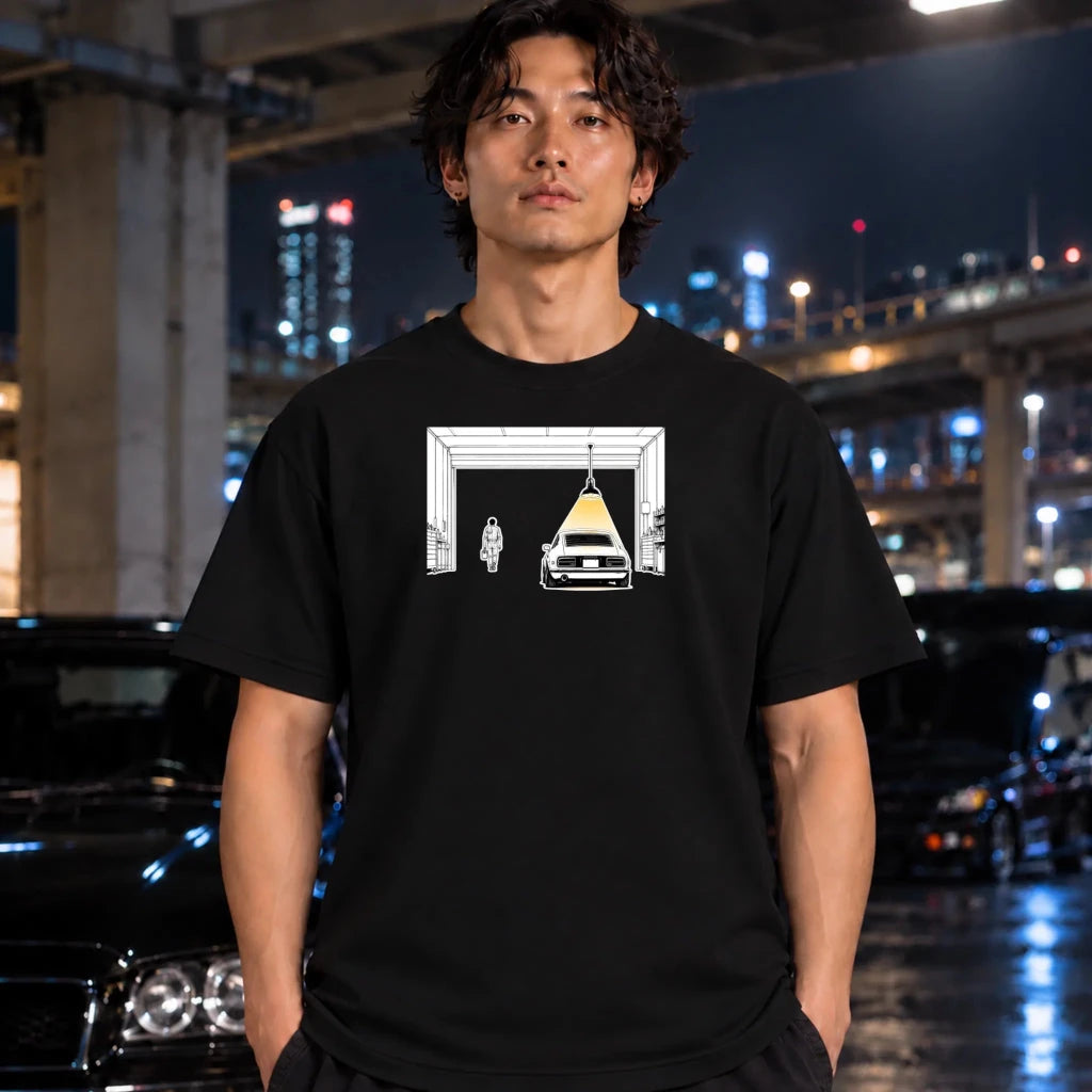 A man wearing a black t-shirt with a graphic print of a car and a figure in front of an open door, standing in a nighttime urban setting with blurred cars and city lights in the background.