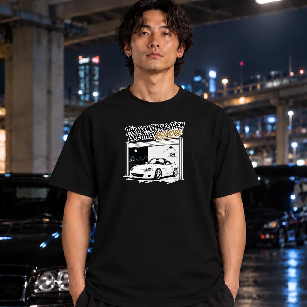 A man wearing a black graphic t-shirt stands in front of a blurred night city background with cars and illuminated buildings. The t-shirt features a white and yellow graphic of a car inside a garage with the text