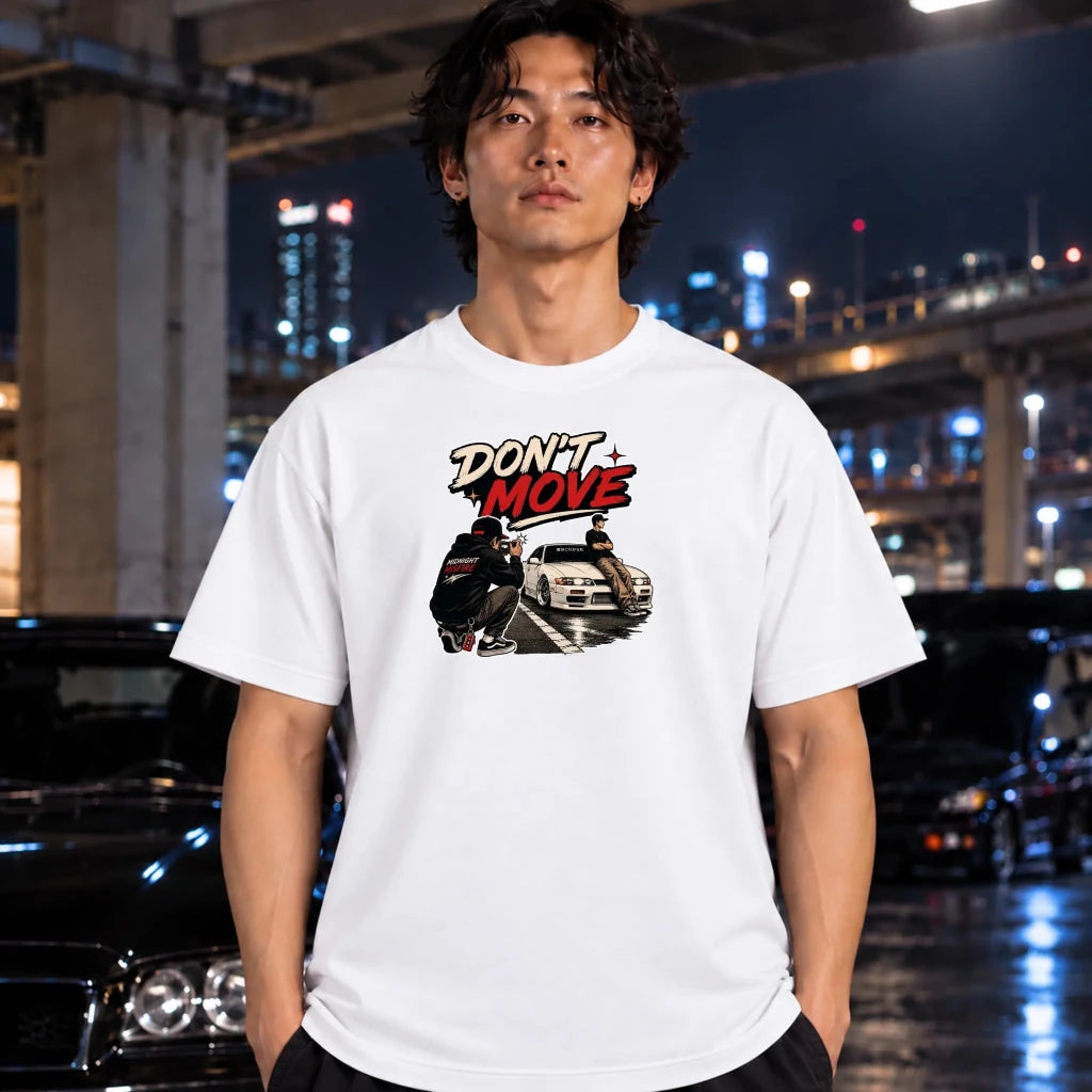 A man wearing a white graphic t-shirt with a 'DON'T MOVE' print featuring a motorcycle and a car, set against a blurred night city background with cars.