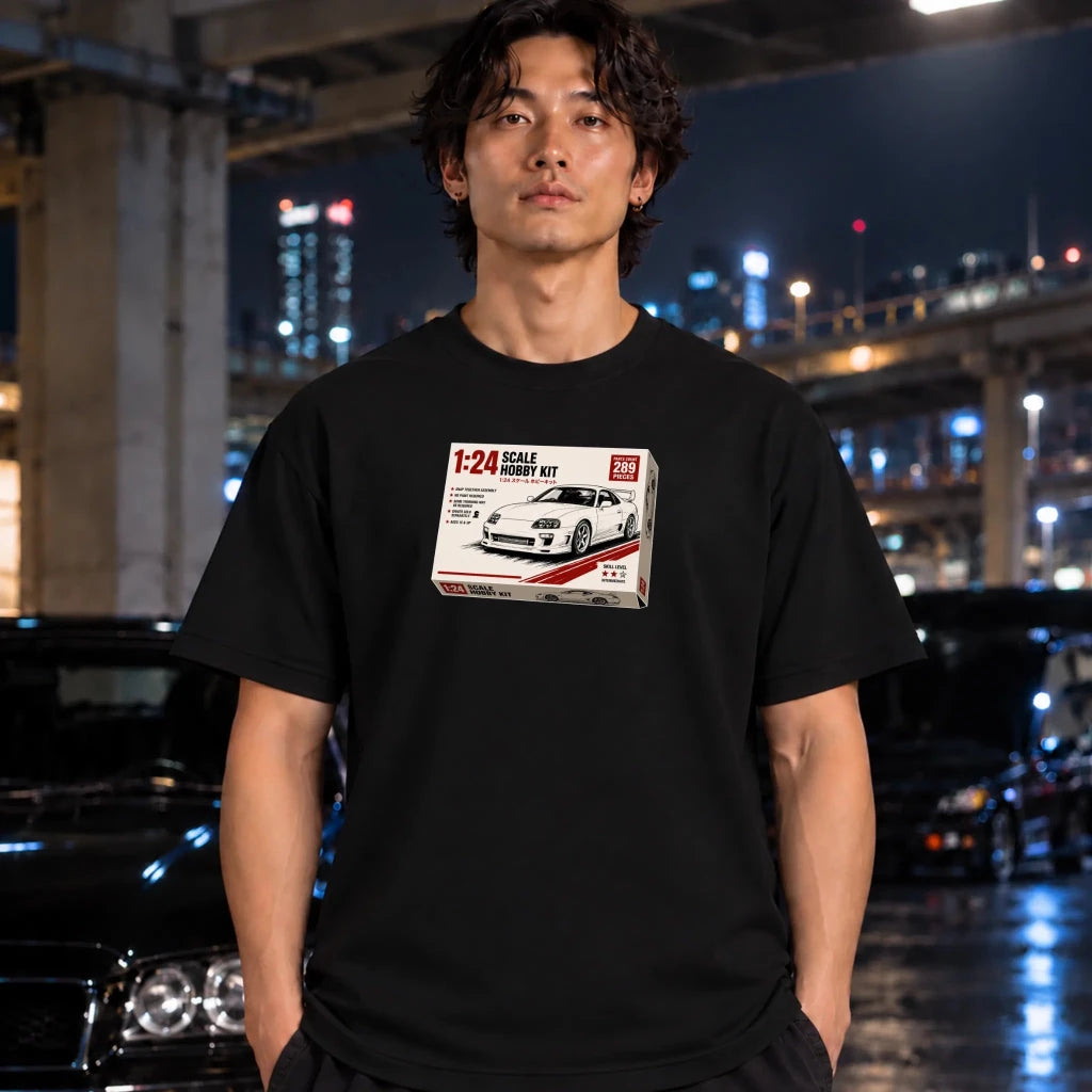 A man wearing a black t-shirt with a printed graphic of a 1:24 scale model kit box, standing in a nighttime urban setting with cars and city lights in the background.