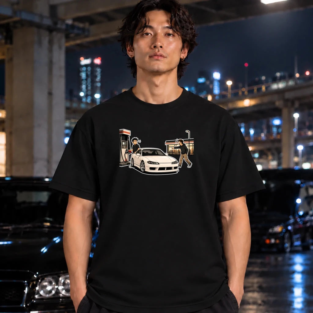 A man wearing a black graphic t-shirt with a white sports car and gas station illustration, standing in a wet urban street at night with blurred cars and city lights in the background.