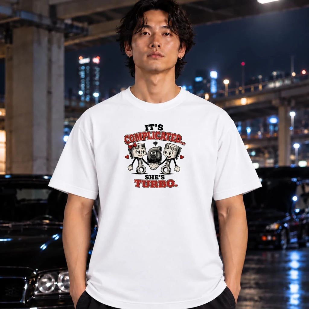 A man wearing a white graphic t-shirt with a turbo and pistons illustration, standing in a wet urban night setting with cars and city lights in the background.