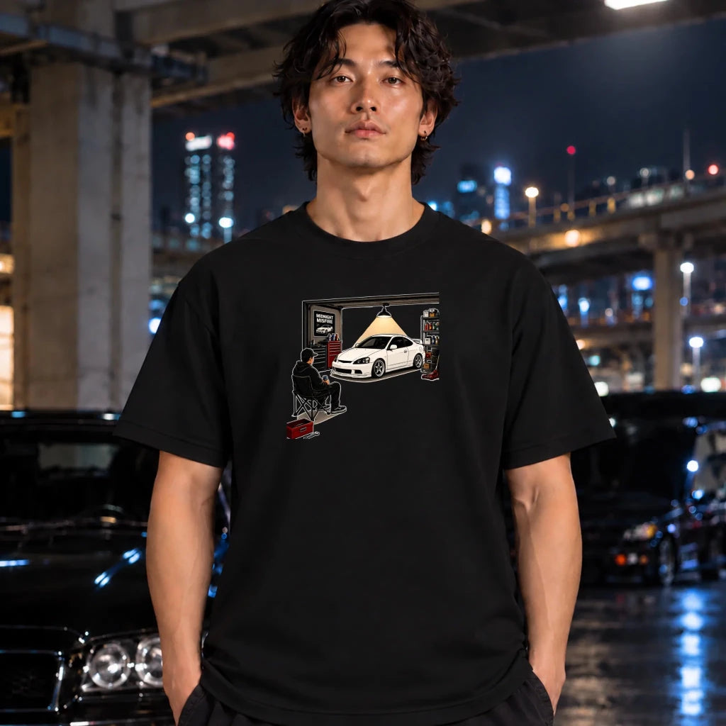 man wearing a black t-shirt featuring a graphic print of a white sports car in a garage with a person sitting in a folding chair. A neon sign and folded dark fabric are partially visible in the corners.