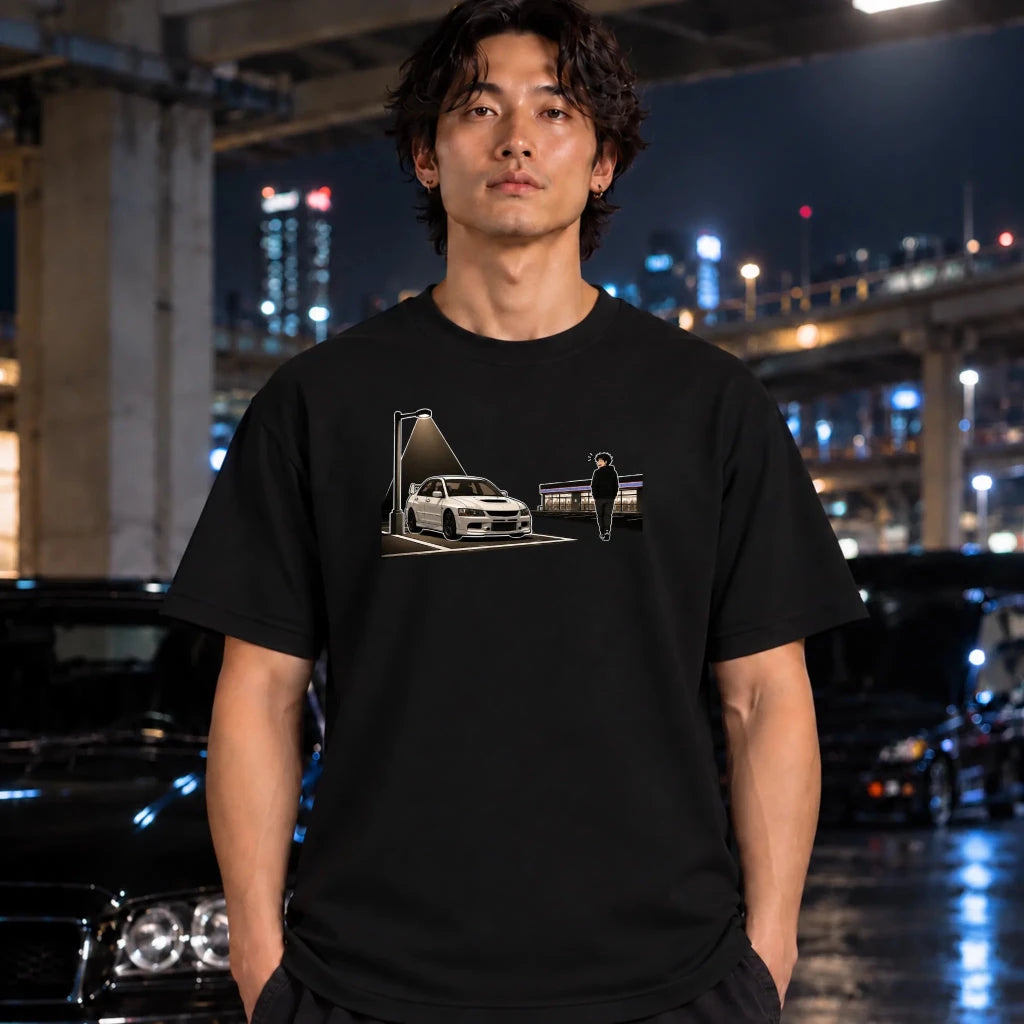 A man wearing a black graphic t-shirt with a white and gray print of a sports car and a figure, standing in a wet urban night scene with cars and city lights in the background.