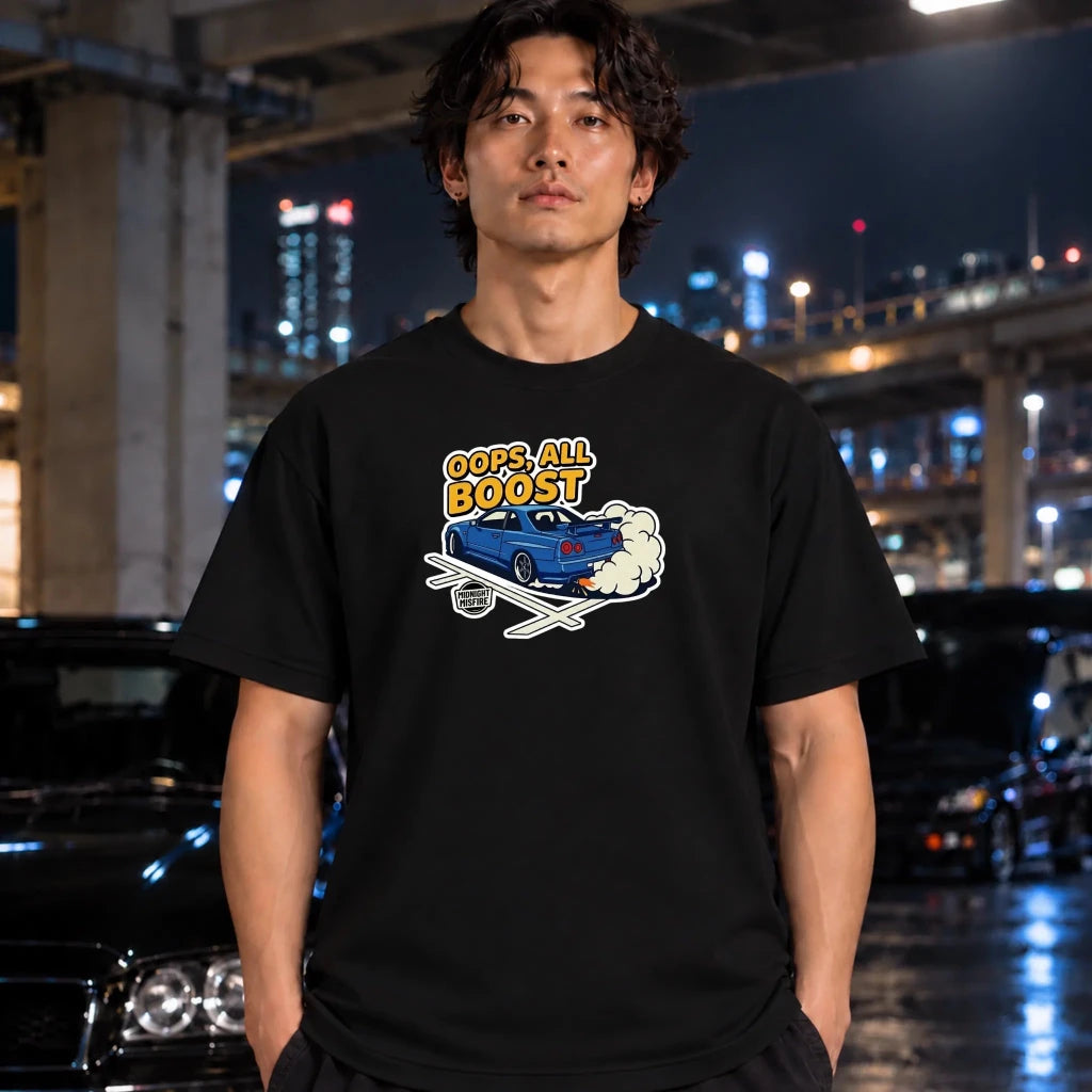 A man wearing a black graphic t-shirt with a blue car and 'OOPS. ALL BOOST' text, standing in front of a blurred night city background with cars.
