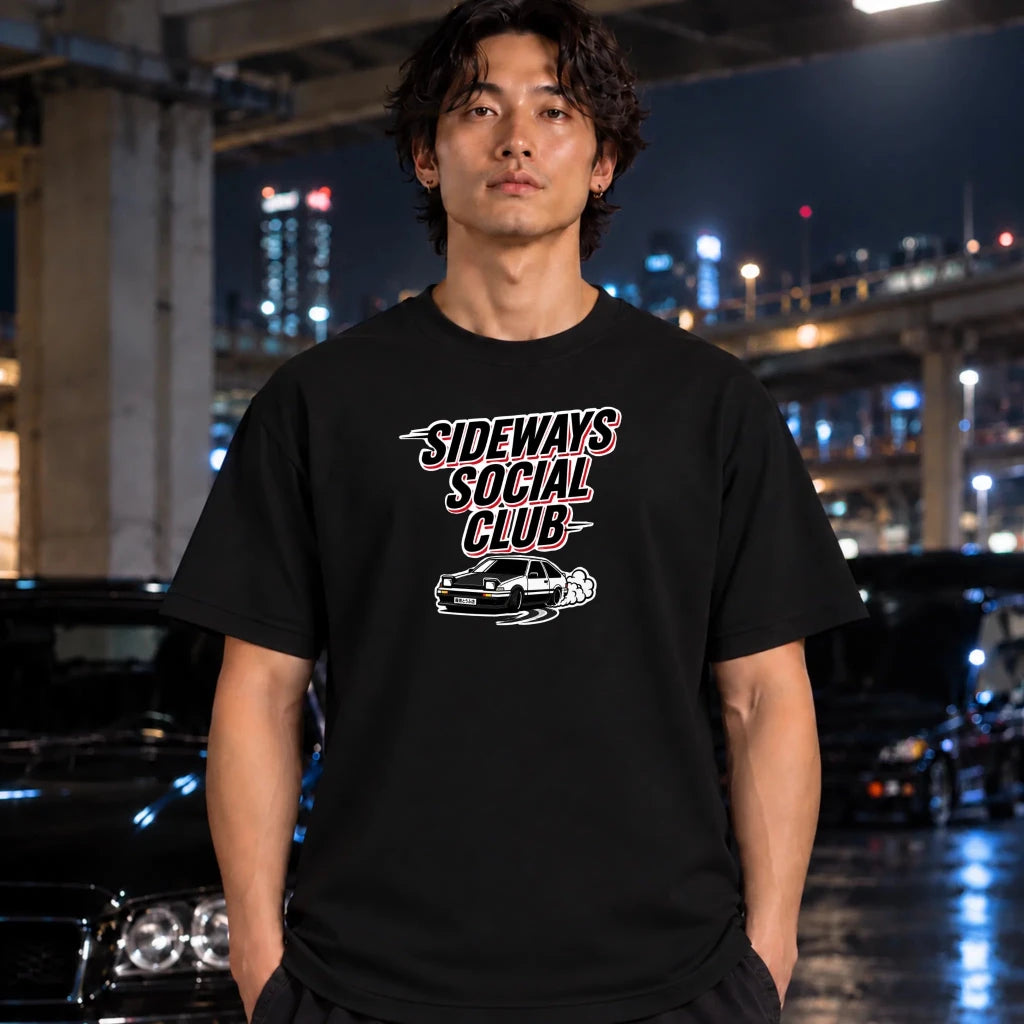 A man wearing a black graphic t-shirt with 'SIDeways SOCIAL CLUB' and a car illustration printed on the chest, standing in a wet urban night scene with cars and city lights in the background.