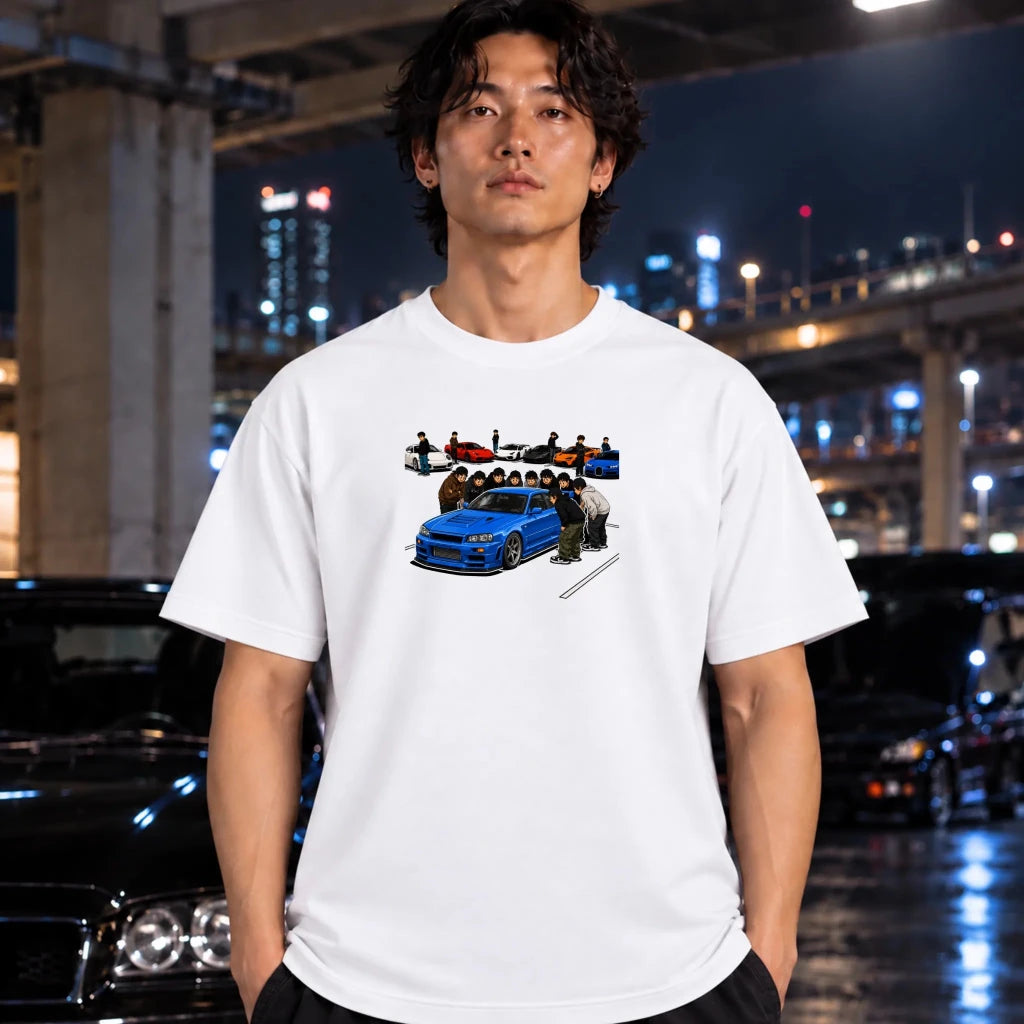 A man wearing a white graphic t-shirt with a blue car and crew illustration, standing in front of a blurred night city background with cars.