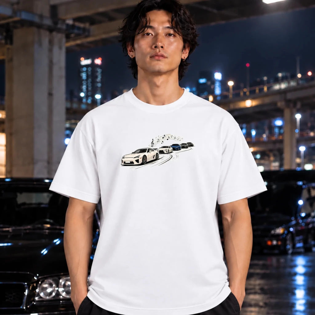 A man wearing a white short-sleeve t-shirt with a graphic of sports cars on the chest, standing in front of a blurred night cityscape with cars and lights.