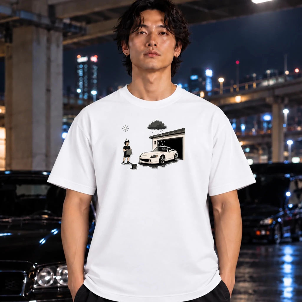 A man wearing a white graphic t-shirt with a black and white illustration of a person standing next to a sports car in front of a garage, set against a blurred night city background with cars and lights.