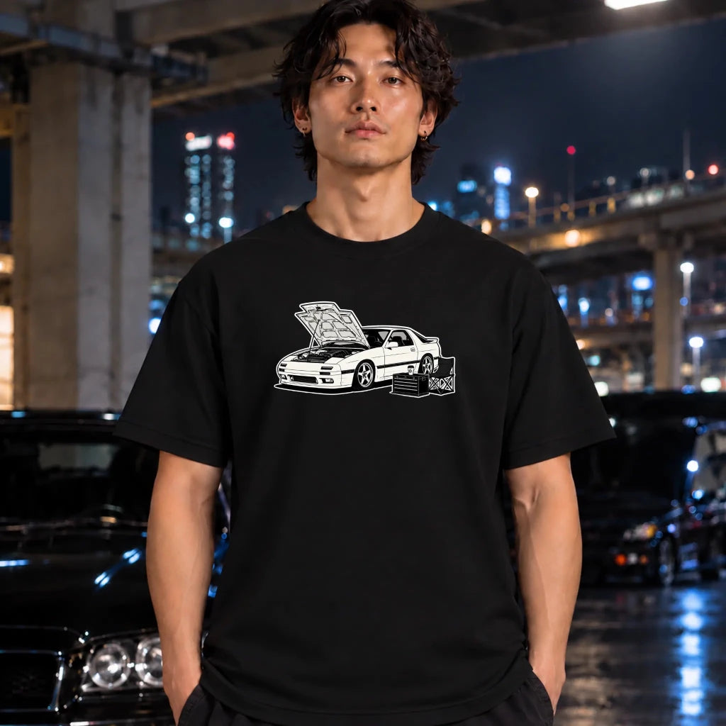 A man wearing a black t-shirt with a white graphic of a sports car standing in front of a blurred night city background with cars.