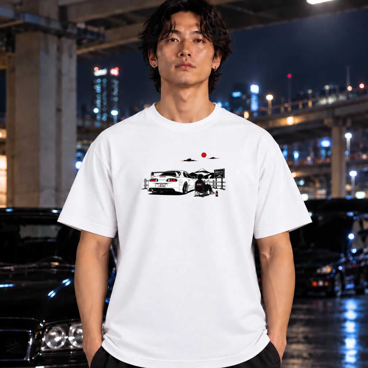 A white short-sleeve graphic T-shirt worn by a man. The shirt features a printed illustration of a white sports car with a red sun and mountain graphic above it.