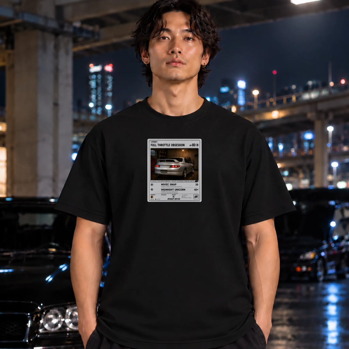 A man wearing a black t-shirt with a graphic print of a silver car standing in a nighttime urban setting with blurred cars and city lights in the background.