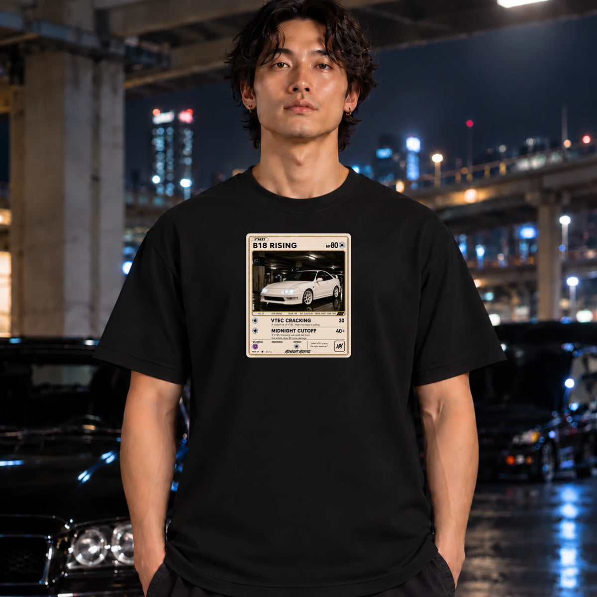 A man wearing a black t-shirt with a vintage-style racing graphic print, standing in a wet urban street at night with blurred cars and city lights in the background.