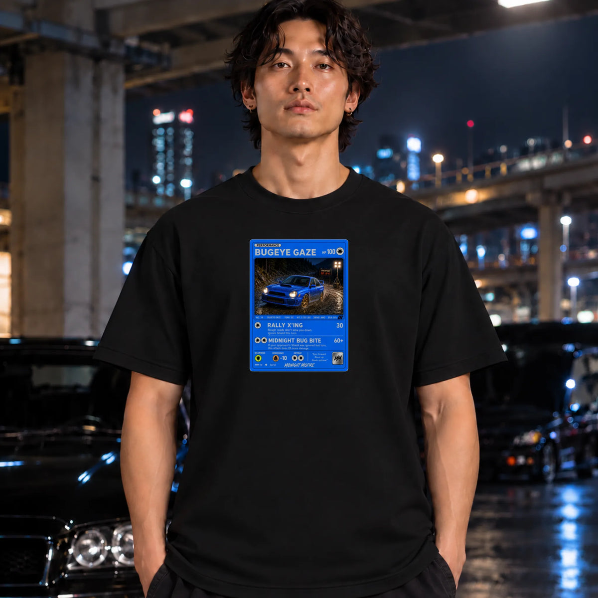 A man wearing a black t-shirt with a blue and white graphic card design on the chest, standing in a nighttime urban setting with blurred city lights and cars in the background.