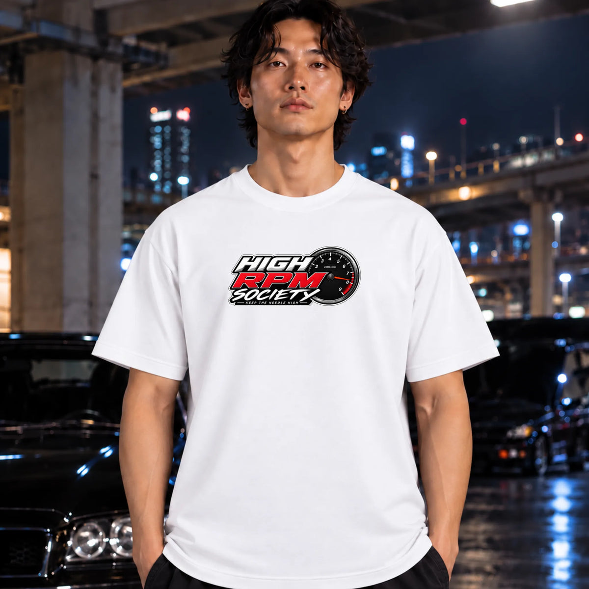 A man wearing a white t-shirt with a 'High RPM Society' graphic printed on the chest, standing in front of a blurred night city background with cars.