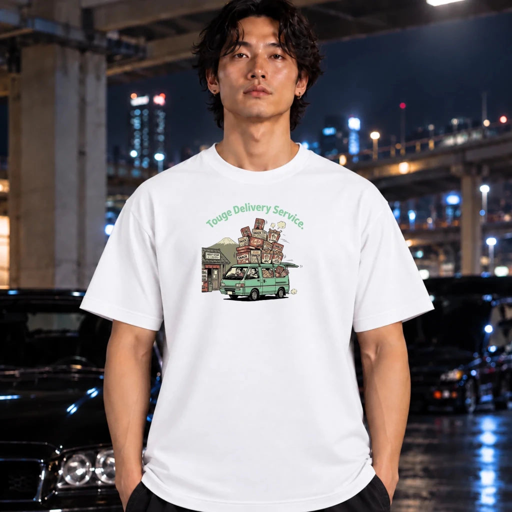 A man wearing a white graphic t-shirt with a 'Touge Delivery Service' van illustration, standing in urban environment with mountain in the background.