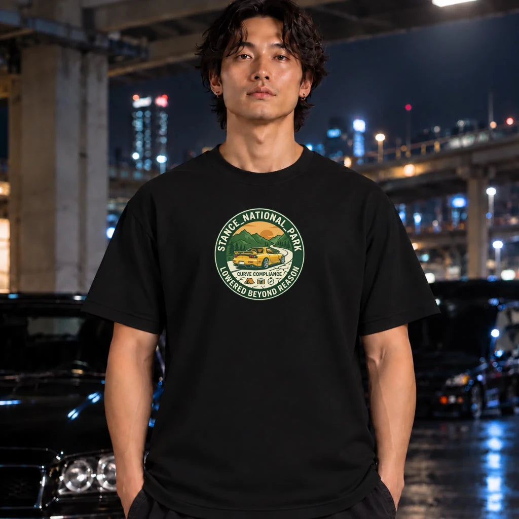 A man wearing a black crewneck t-shirt with a circular green and yellow 'Stance National Park' graphic on the chest, standing in a wet urban night setting with cars and city lights in the background.