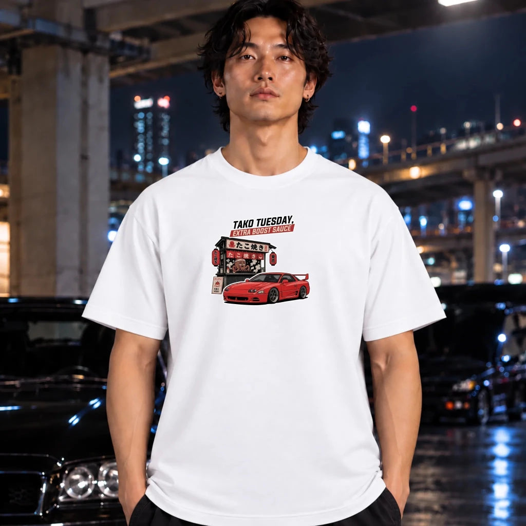 A man wearing a white graphic t-shirt with a red sports car and 'TAKO TUESDAY' text, standing in front of a blurred night city background with cars.