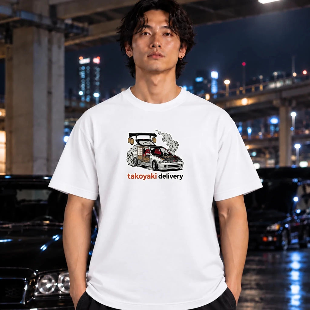 A man wearing a white graphic t-shirt with a cartoon car and the text 'takoyaki delivery' printed on the chest, standing in front of a blurred night city background with cars.