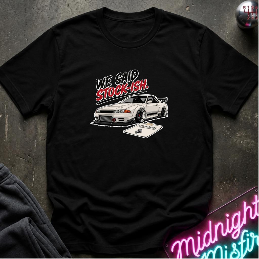 A black short-sleeve graphic t-shirt laid flat on a dark textured surface. The shirt features a white sports car illustration with the text 'WE SAID STOCK-ISH!' in white and red.