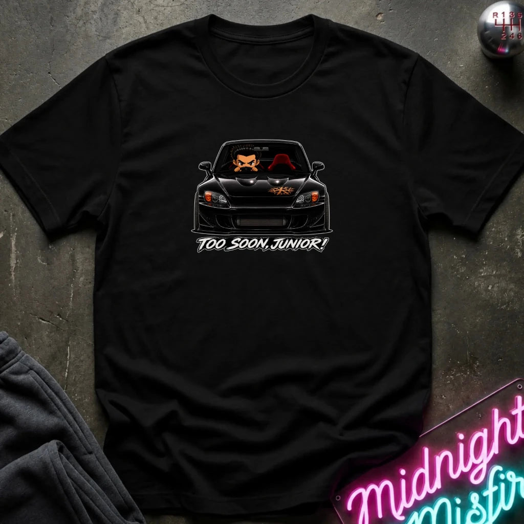 A black short-sleeve t-shirt laid flat on a dark textured surface, featuring a graphic of a black sports car with orange and red details and the text 'TOO SOON, JUNIOR!' printed below it.