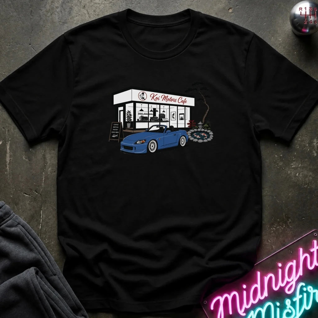 A black  short-sleeve t-shirt laid flat on a dark textured surface, featuring a graphic print of a blue sports car parked in front of 'Koi Motors Cafe' with a small tree illustration. 