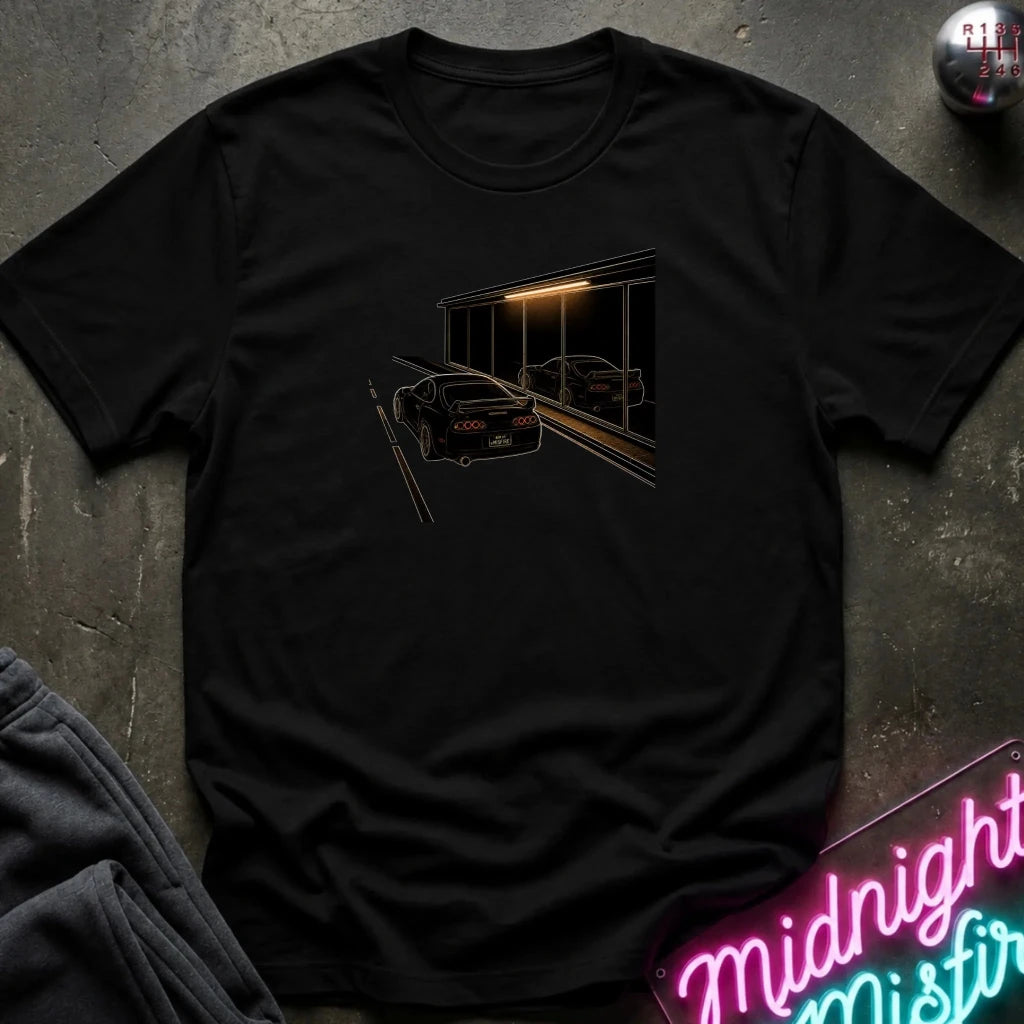 A black short-sleeve t-shirt laid flat on a dark textured surface, featuring a graphic print of a black sports car in front of a dark building with warm light. 
