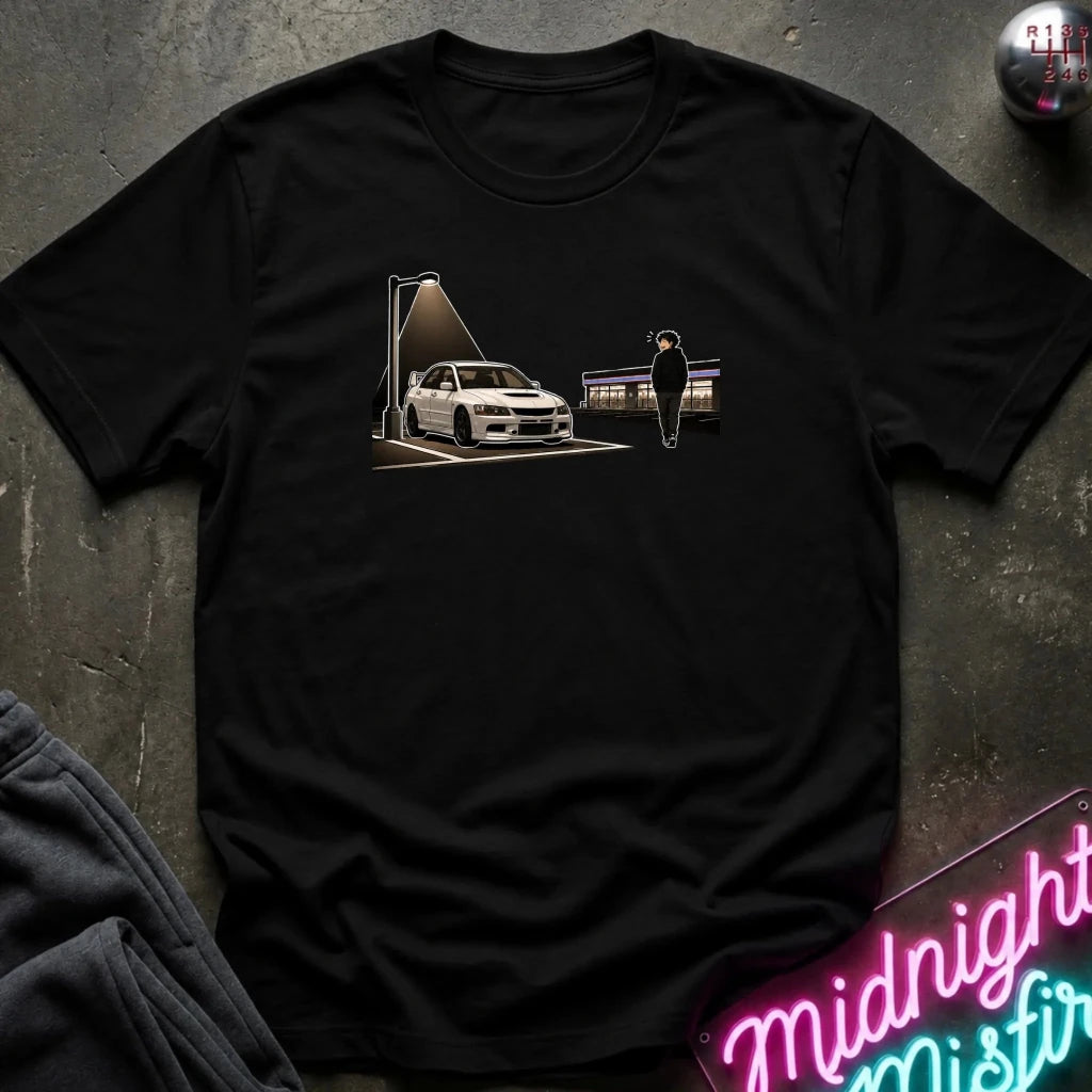 A black short-sleeve crewneck t-shirt laid flat on a dark textured surface. The shirt features a graphic print of a white sports car under a spotlight with a silhouette figure standing nearby.
