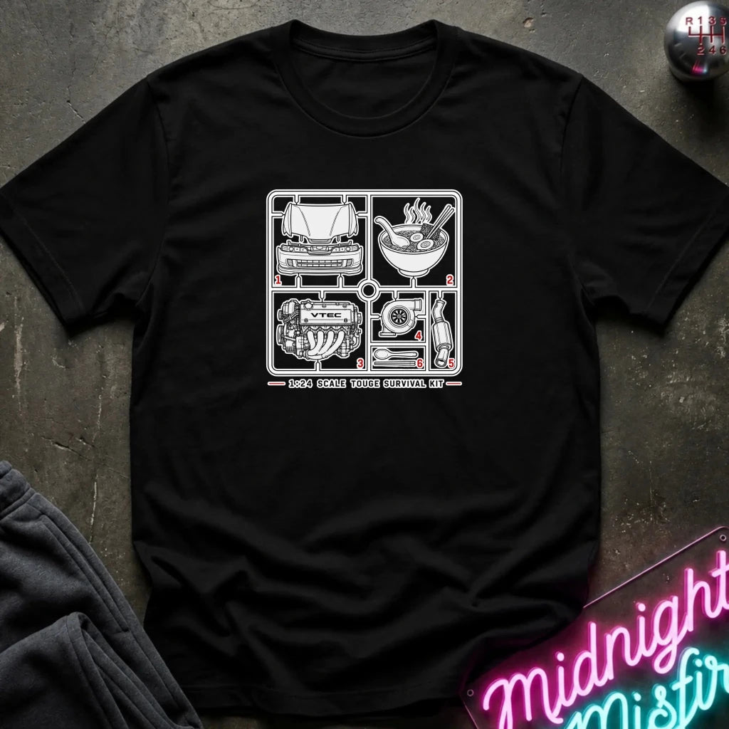 A black short-sleeve crewneck t-shirt laid flat on a dark textured surface. The shirt features a white graphic print in the center depicting automotive and culinary elements (a grill, a bowl of ramen, an engine, and a bottle) 