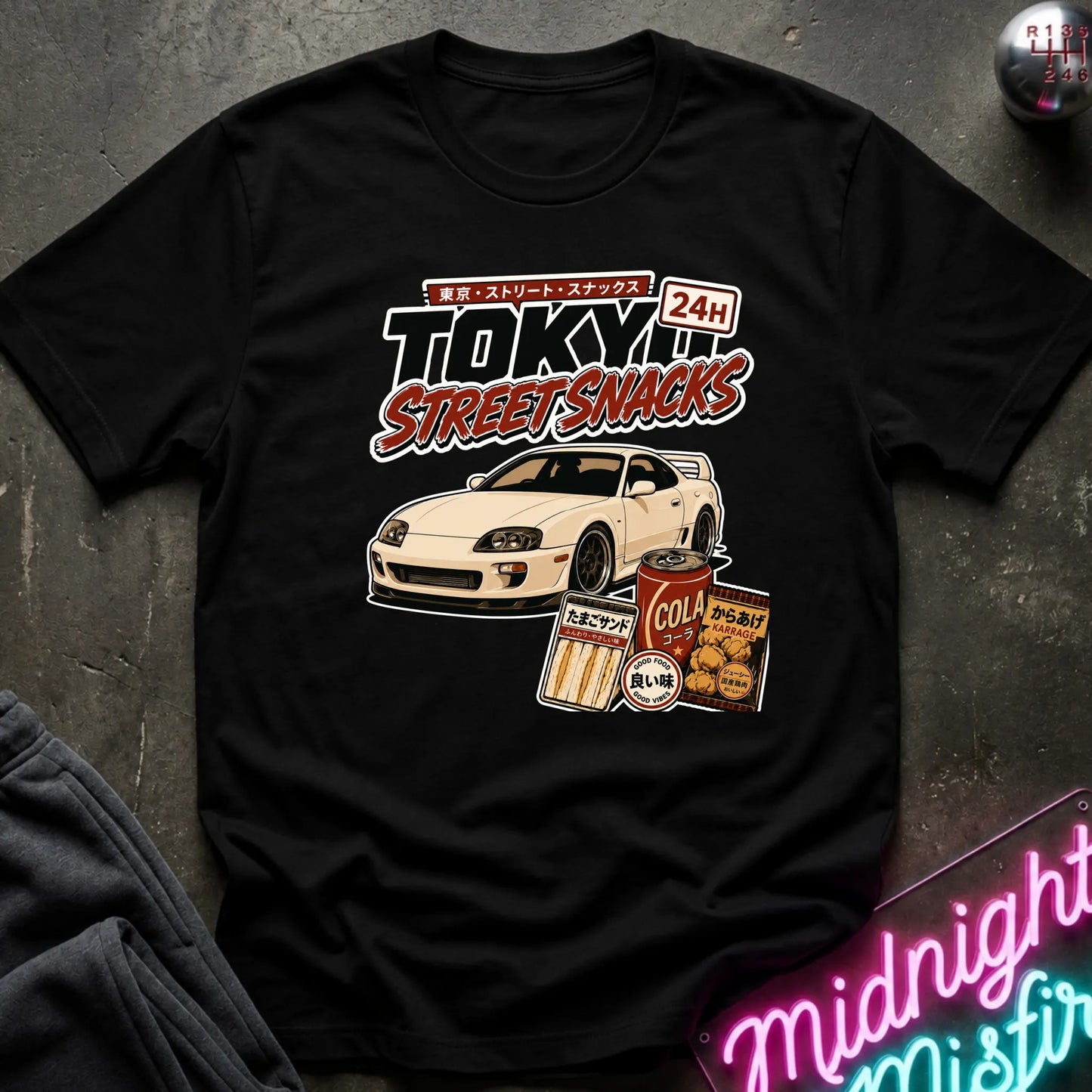 A black graphic t-shirt laid flat on a dark textured surface. The graphic features a white sports car and retro Japanese-style snack cans with the text 'TOKYO STREET SNACKS 24H'.
