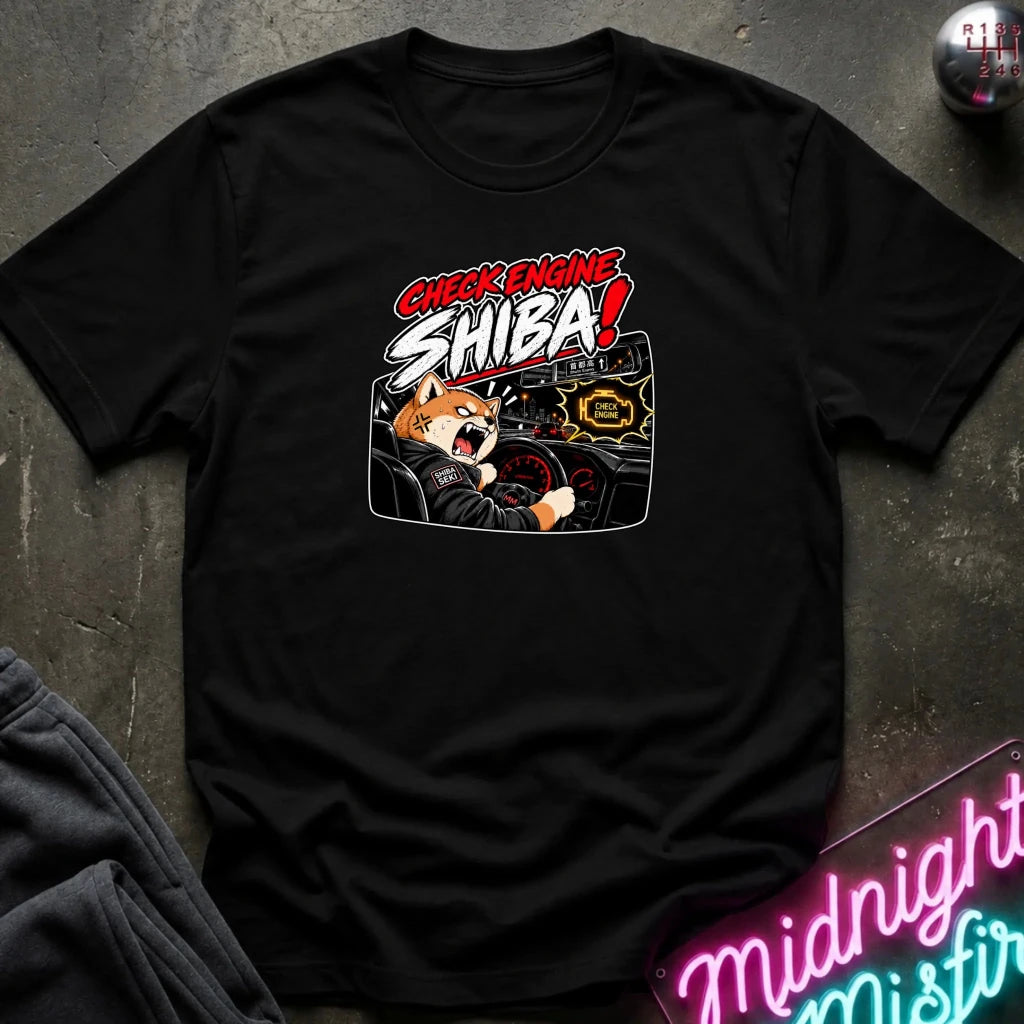 A black short-sleeve graphic t-shirt laid flat on a dark textured surface. The graphic features the text 'CHECK ENGINE SHIBA!' in red and white, above an illustration of an angry Shiba Inu dog behind a car dashboard.