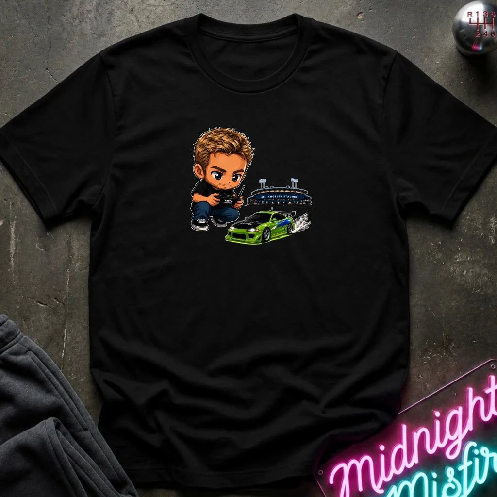 A black graphic t-shirt laid flat on a dark textured surface. The graphic features a green car, and person  controlling it with remote.