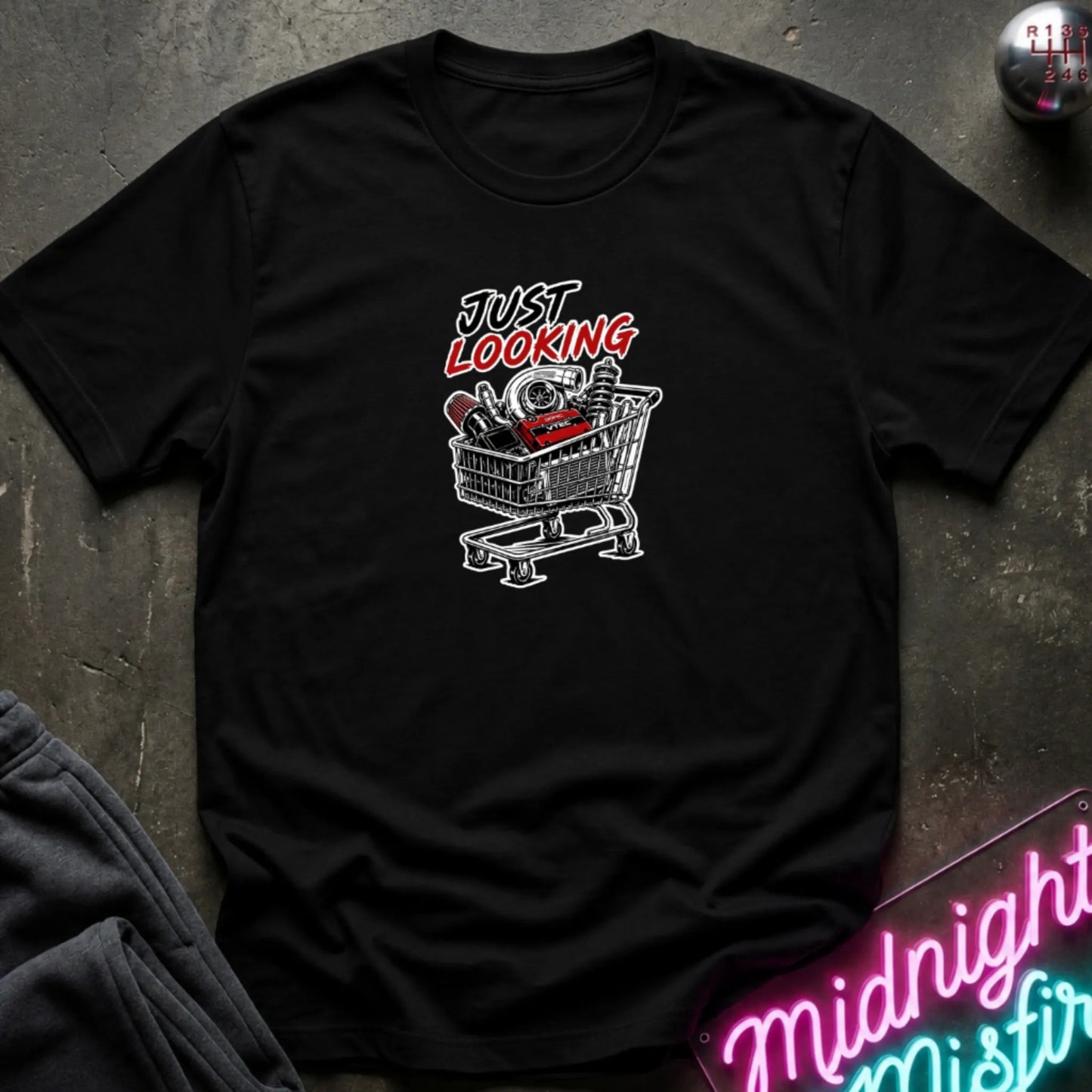 A black short-sleeve t-shirt laid flat on a dark textured surface, featuring a graphic print of a shopping cart filled with items and the text 'JUST LOOKING' in black and red. Partially visible props include dark pants in the lower left and a neon-style sign in the lower right.