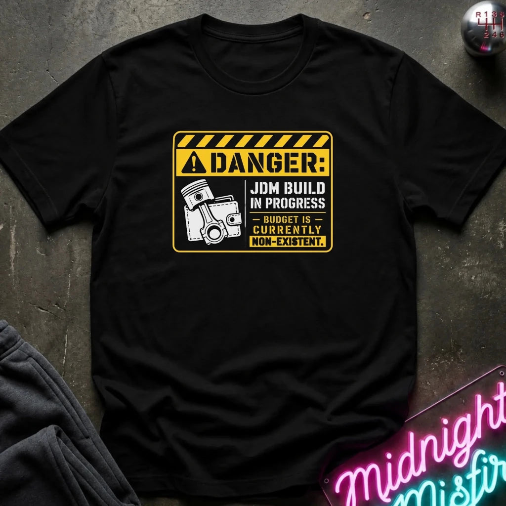 A black short-sleeve graphic T-shirt laid flat on a dark textured surface. The shirt features a yellow and black warning-sign style print with the text 'DANGER: JDM BUILD IN PROGRESS BUDGET IS CURRENTLY NON-EXISTENT.