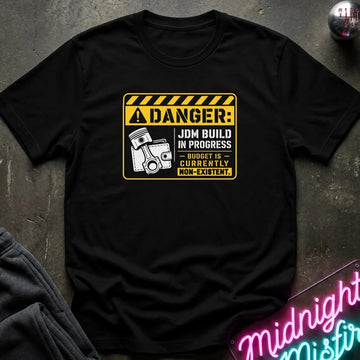 A black short-sleeve graphic T-shirt laid flat on a dark textured surface. The shirt features a yellow and black warning-sign style print with the text 'DANGER: JDM BUILD IN PROGRESS BUDGET IS CURRENTLY NON-EXISTENT.