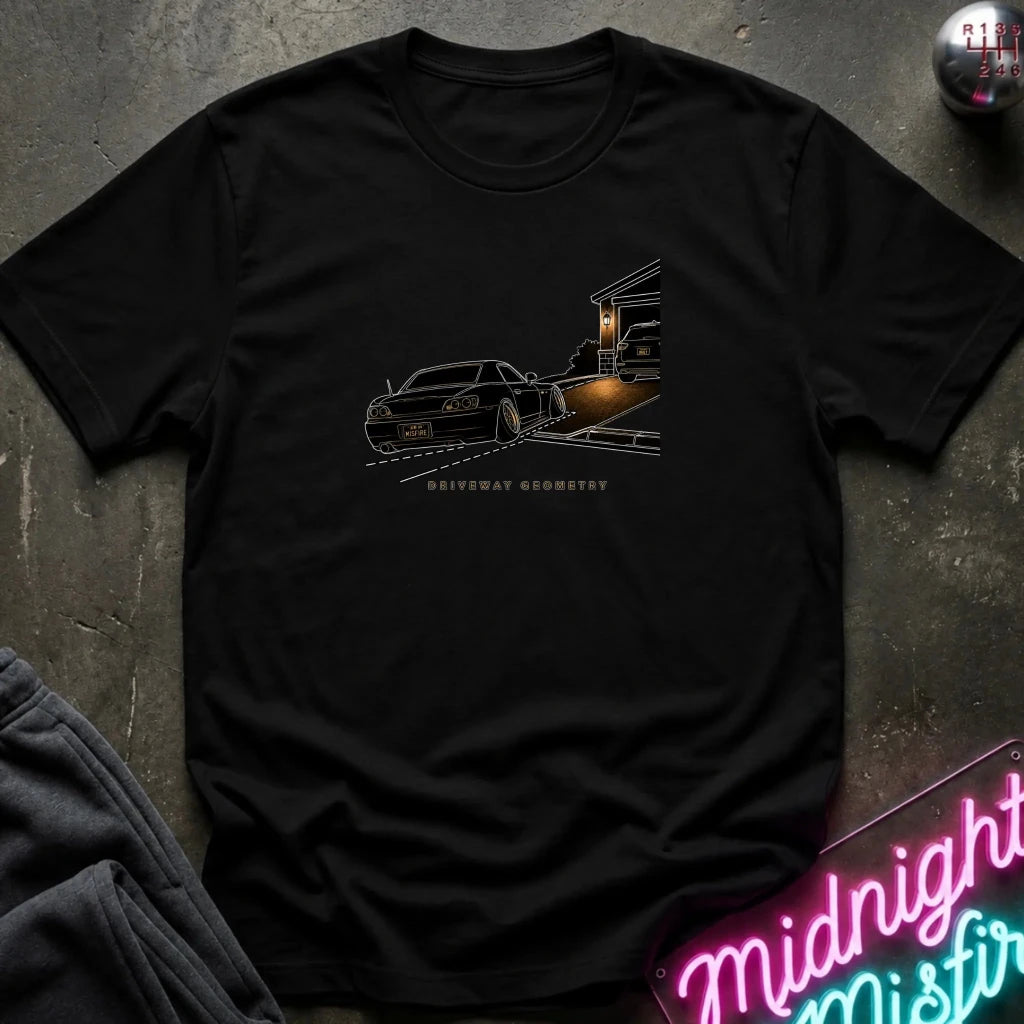 A black short-sleeve t-shirt laid flat on a dark textured surface, featuring a graphic print of a black classic car going up the driveway.