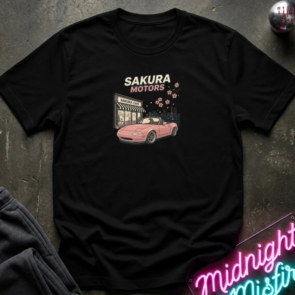 A black short-sleeve graphic t-shirt laid flat on a dark textured surface. The graphic features the text 'SAKURA MOTORS' above an illustration of a pink convertible car parked in front of a convenience store, with cherry blossoms falling.