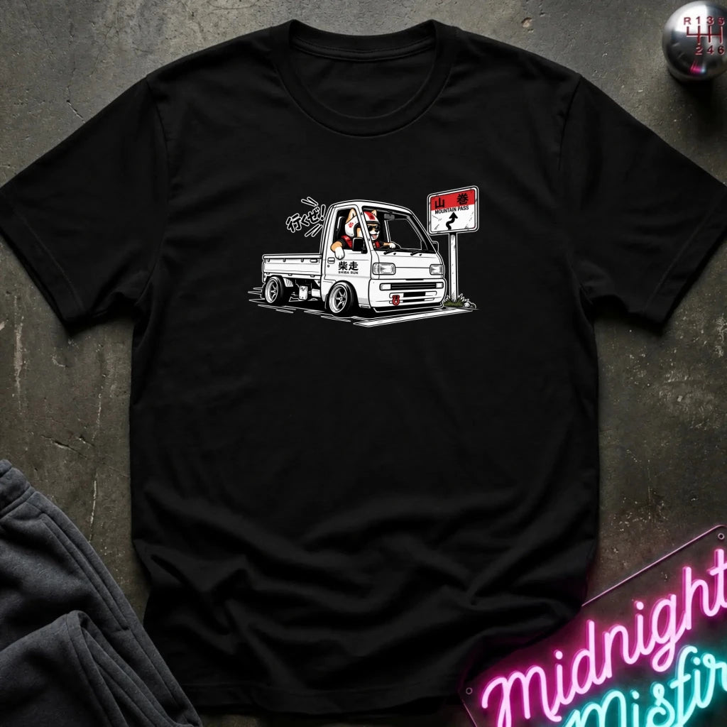 A black short-sleeve crewneck t-shirt featuring a graphic of a white Japanese pickup truck with a dog driver and a sign, laid flat on a dark textured surface.