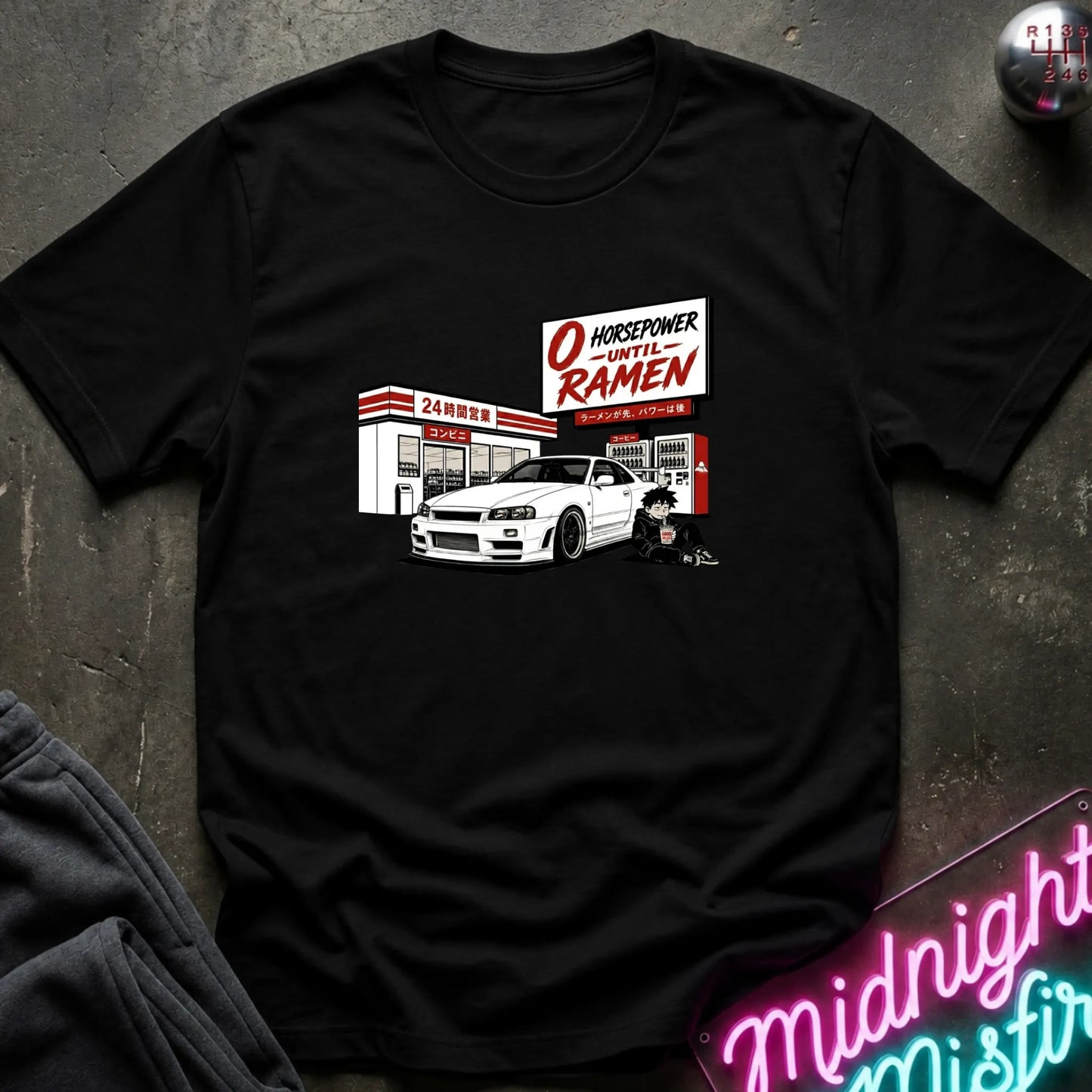 A black short-sleeve graphic t-shirt laid flat on a dark textured surface. The graphic features a white sports car parked in front of a store  with a sign reading 'O HONORPOWER -UNTIL RAMEN'.