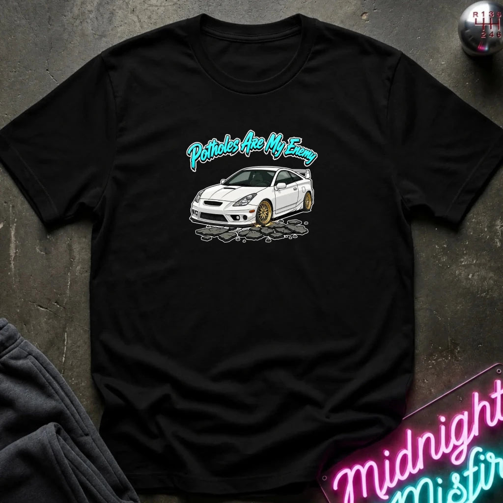 A black short-sleeve graphic t-shirt laid flat on a dark textured surface. The shirt features a white sports car illustration with gold wheels and the text 'Potholes Are My Enemy' in a teal script above it.