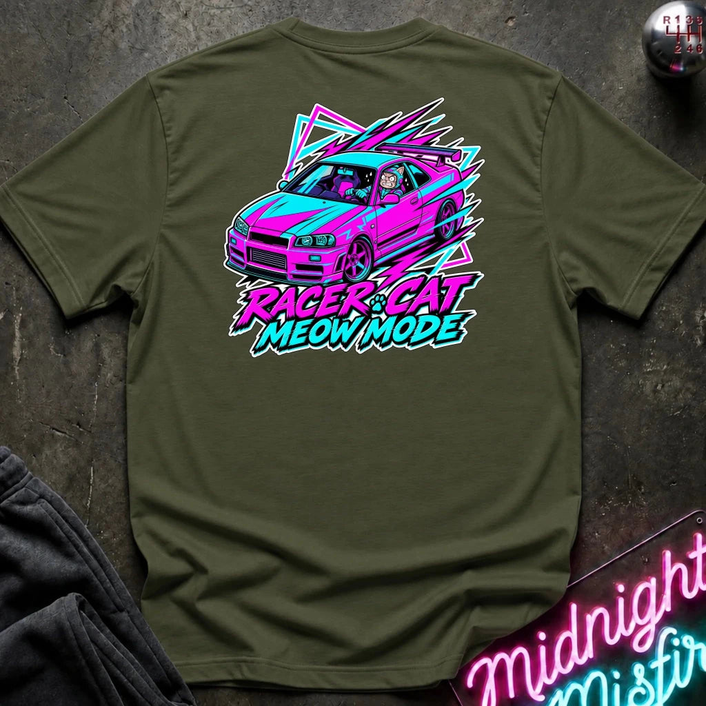 An olive green t-shirt laid flat on a dark textured surface, featuring a colorful graphic of a pink and blue race car with the text 'RACER CAT MEOW MODE' in neon pink and blue. A neon sign and folded dark fabric are partially visible in the background.