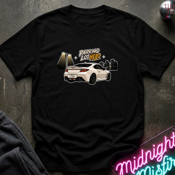 A black short-sleeve graphic t-shirt laid flat on a dark textured surface. The graphic features a white sports car with the text 'PARKING LOT HERO' in white and orange, alongside a gold-toned gate and a dark city skyline.