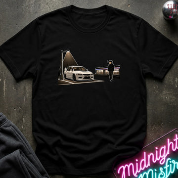 A black short-sleeve crewneck t-shirt laid flat on a dark textured surface. The shirt features a graphic print of a white sports car under a street lamp with a silhouette of a person standing nearby.