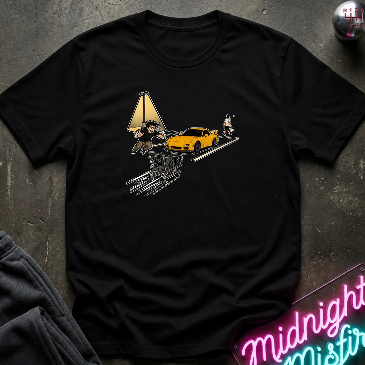 A black short-sleeve graphic t-shirt laid flat on a dark textured surface. The graphic shows a yellow sports car on a road with a shopping cart.