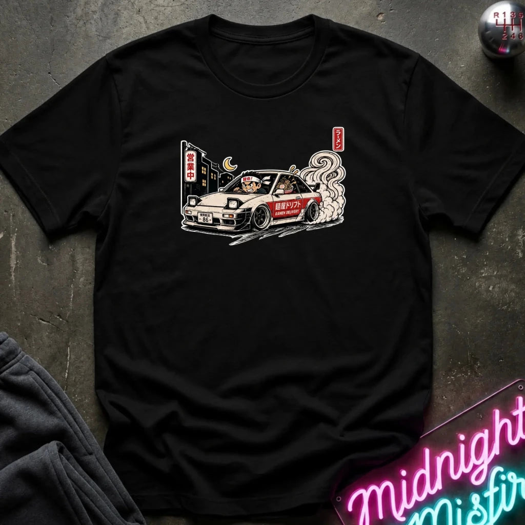 A black short-sleeve graphic t-shirt laid flat on a dark textured surface. The shirt features a detailed illustration of a white sports car with Japanese text and smoke, styled with a neon sign and dark clothing in the background.