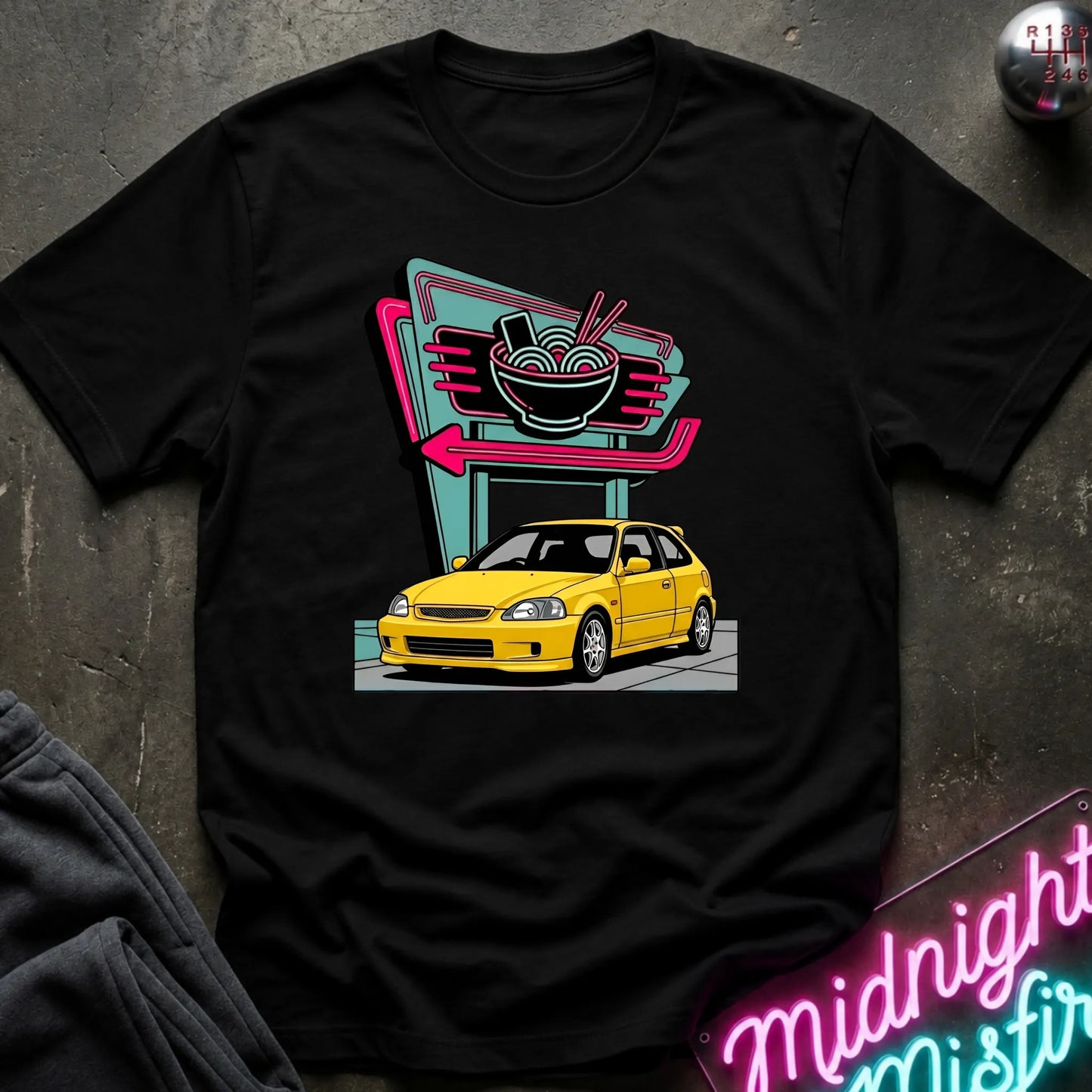 A black short-sleeve graphic t-shirt laid flat on a dark textured surface. The shirt features a colorful retro-style print of a yellow car parked in front of a neon ramen shop sign.