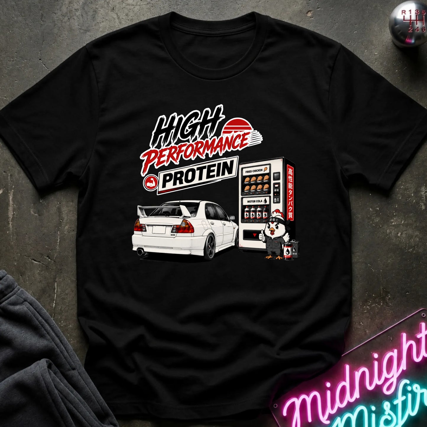 A black short-sleeve graphic T-shirt laid flat on a dark textured surface. The shirt features a large front print with the words “High Performance Protein,” a white car, a vending machine illustration, and a small character.