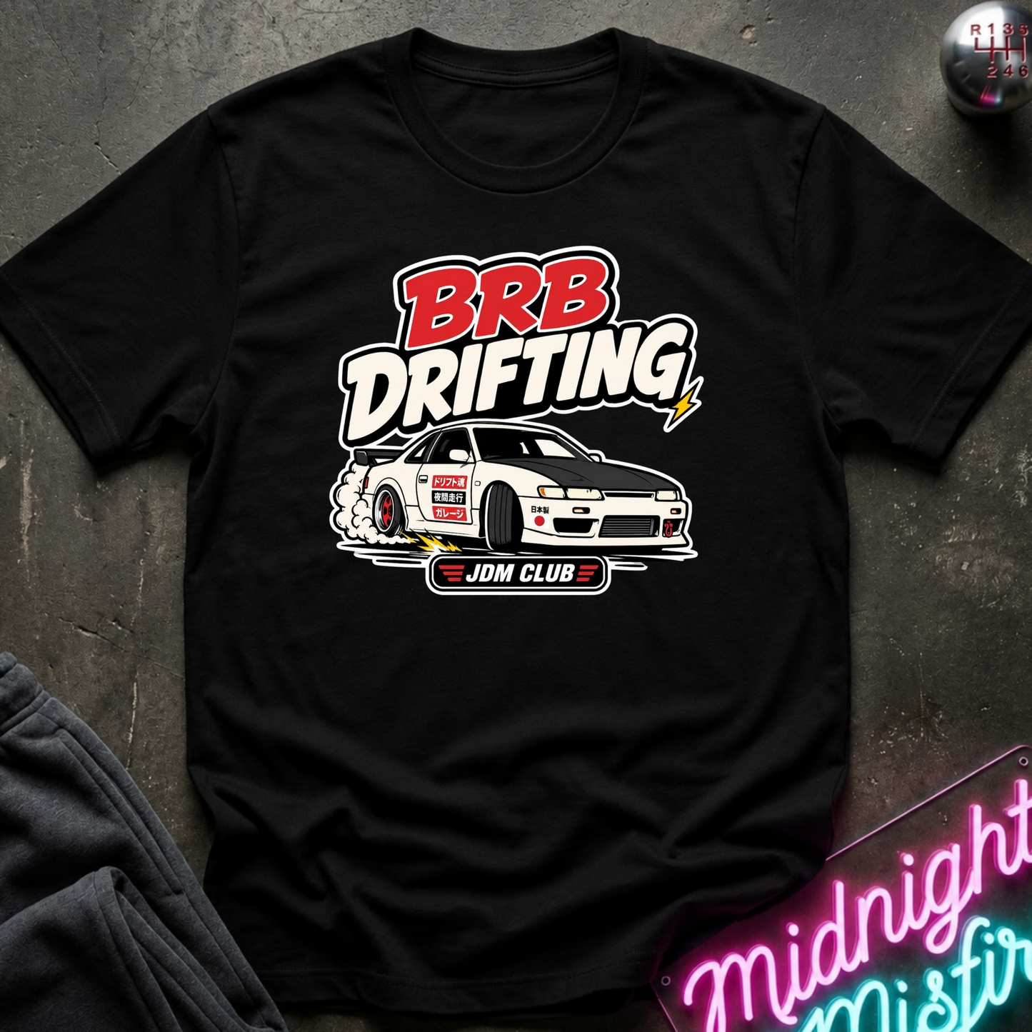A black short-sleeve graphic T-shirt laid flat on a dark textured surface. The shirt features a large front print with the words “BRB DRIFTING” in red and white, a white drifting car illustration, and “JDM CLUB” below.
