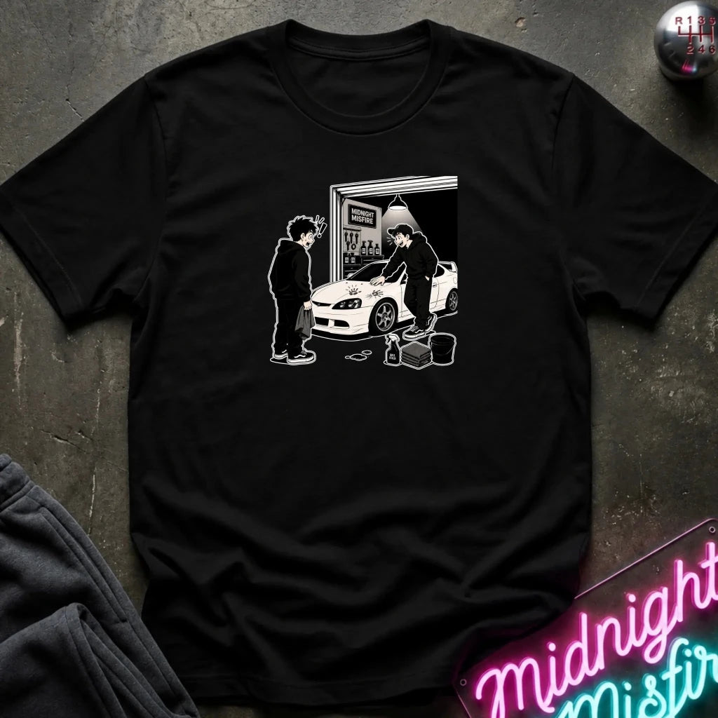A black short-sleeve crewneck t-shirt with a white graphic print of two people standing by a white sports car in a garage. The shirt is laid flat on a dark textured surface with a neon sign and folded gray fabric visible in the corners.