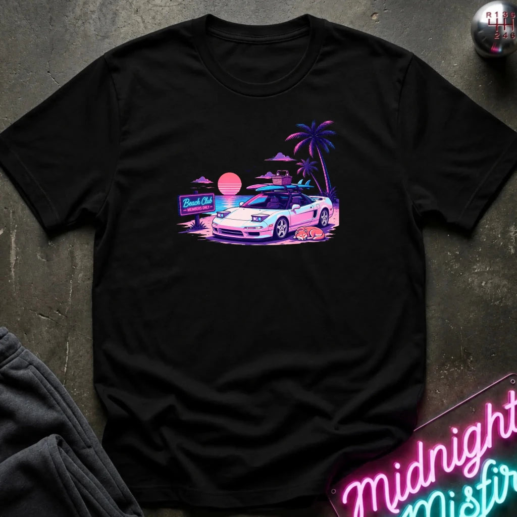 A black short-sleeve t-shirt laid flat on a dark textured surface, featuring a colorful retro graphic of a white sports car on a beach with palm trees and a sunset.